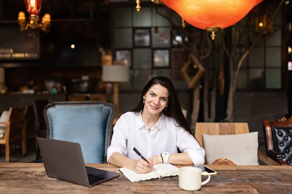 Joven mujer de negocios caucásica con el pelo largo y morena trabajando en el ordenador portátil en la cafetería. Estudiante universitario que usa tecnología, educación en línea, freelance — Foto de Stock