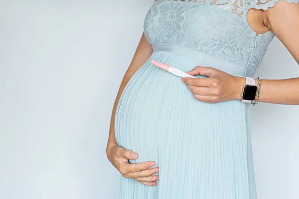 Närbild fotografi av ung kvinna som håller positiva gravida test vidrör hand hennes stora mage i blå klänning Isolerad på vit bakgrund med kopieringsutrymme. Hudvård med återfuktande kräm mot — Stockfoto