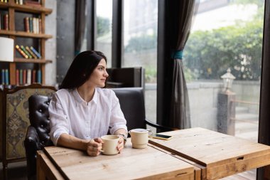 Uzun esmer saçlı, günlük kıyafetler giyen ve kafeteryada elinde kahve fincanı olan genç beyaz bir kadın. Hayatı ya da işi düşünürken çay içen orta yaşlı bir kadın.