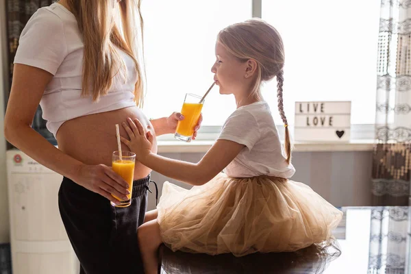 Ciężarna matka i córka piją świeży sok z mango w kuchni. Życie rodzinne podczas covid-19 kwarantanny koronawirusowej w domu, izolacja, koncepcja zdrowego stylu życia. — Zdjęcie stockowe