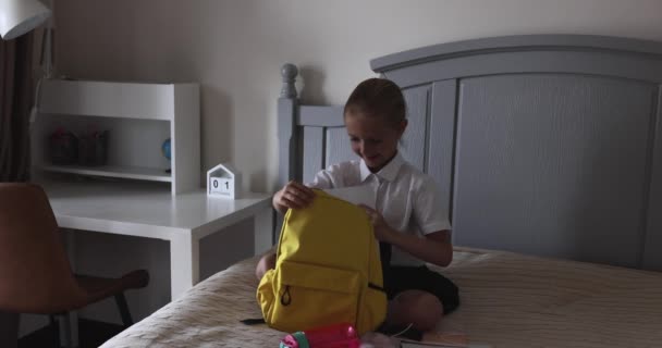 Мила біла дівчина початкової школи в уніформі і упаковці рюкзака вдома на ліжку, дитина готується до першого дня в школі після закінчення коронавірусу ковірусу-19 карантину. Повільний рух — стокове відео