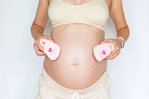 Fotografi closeup dari wanita muda memegang sepatu merah muda kecil untuk bayi perempuan dan menyentuh tangannya perut besar telanjang terisolasi di latar belakang putih dengan salinan ruang. Kulit peduli dengan krim pelembab — Stok Foto