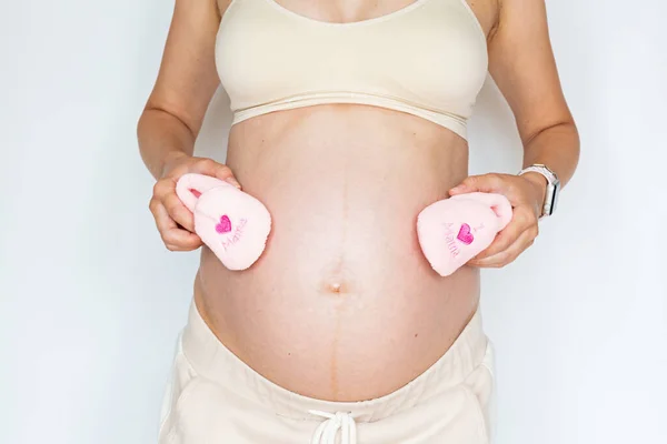 Fotografía de cerca de la joven mujer sosteniendo pequeños zapatos rosados para la hija del bebé y tocando las manos de su gran vientre desnudo aislado sobre fondo blanco con espacio de copia. Cuidado de la piel con crema hidratante —  Fotos de Stock