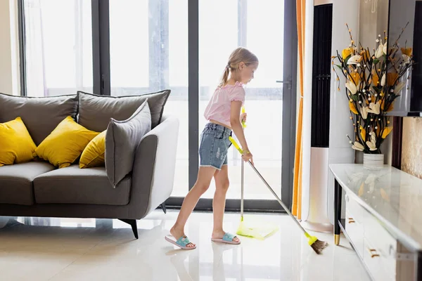 Little Caucasian blonde girl with blonde hair seven years old cleaning floor in living room. Modern home interior, domestic life. — Stock Photo, Image