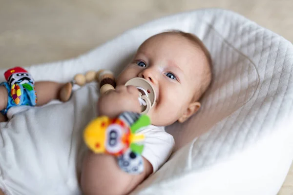 Bonito bebê loiro caucasiano três meses de idade deitado em casulo em casa. Criança vestindo nas mãos e pernas recheado brinquedo som educacional, mamilo chupando. — Fotografia de Stock