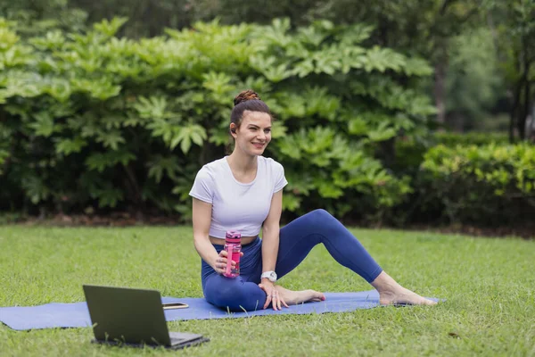 Mujer caucásica joven haciendo ejercicio en línea en el parque de verano durante la pandemia del coronavirus COVID-19. Distanciamiento social durante el entrenamiento físico. Chica que usa ropa deportiva al estirar y practicar yoga — Foto de Stock