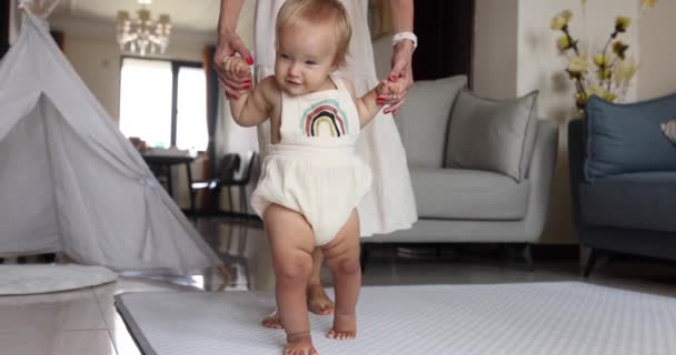 Niedliche kaukasische Baby Mädchen mit blonden Haaren und blauen Augen ein Jahr alt lernen zu gehen. Säugling macht erste Schritte zu Hause. Mutter unterstützt Kind beim Gehen Glückliches neues Lebenskonzept. Kleine Füße — Stockvideo