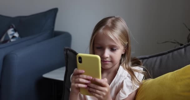 快乐的白种人小女孩，7岁的金发，在家里沙发上用智能手机。可爱的孩子有乐趣与手机游戏和使用应用程序。4k慢动作 — 图库视频影像