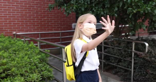 Glad liten kaukasiska blond flicka sju år gammal i uniform med gul ryggsäck står nära skolan i ansiktsmask efter sommarsemester eller Coronavirus covid-19 karantän. 4k slow motion — Stockvideo