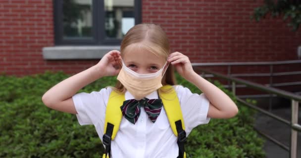 Glückliches kleines kaukasisches blondes Mädchen von sieben Jahren in Uniform mit gelbem Rucksack, das nach den Sommerferien in der Nähe der Schule mit Gesichtsmaske oder Coronavirus covid-19-Quarantäne steht. 4k Zeitlupe — Stockvideo