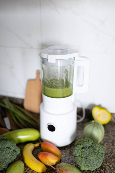 Fresh ingredients for healthy detox cocktail green color. Blender with smoothie for diet on kitchen table, next lying broccoli and other vegetables and fruits.