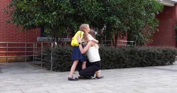 Glad liten kaukasiska blond flicka sju år gammal i uniform med gul ryggsäck går tillbaka till skolan efter sommarsemester eller Coronavirus covid-19 karantän med ung mor. 4k slow motion — Stockvideo