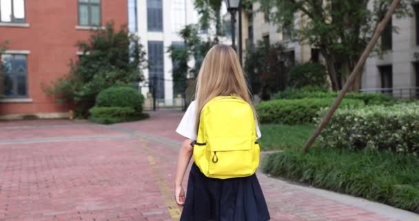 Glad liten kaukasiska blond flicka sju år gammal i uniform med gul ryggsäck går tillbaka till skolan efter sommarsemester eller Coronavirus covid-19 karantän. 4k slow motion — Stockvideo