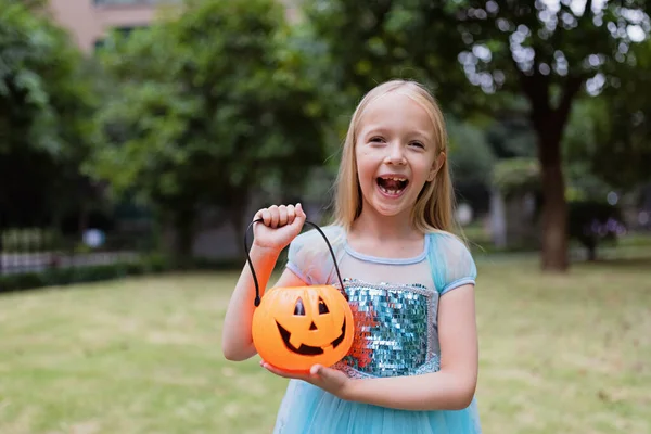 Lilla blonda flicka i prinsessdräkt med pumpa lykta i parken. Glad Halloween koncept — Stockfoto