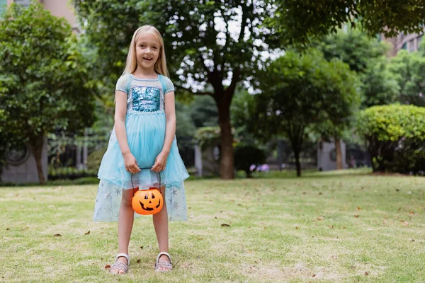 Mała blondynka w kostiumie księżniczki z dyniową latarnią w parku. Szczęśliwego Halloween koncepcja — Zdjęcie stockowe