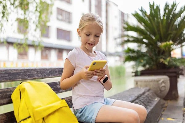 Skolflicka tillbaka till skolan efter sommarlovet. Lyckligt barn med hjälp av mobil phome utomhus på morgonen — Stockfoto