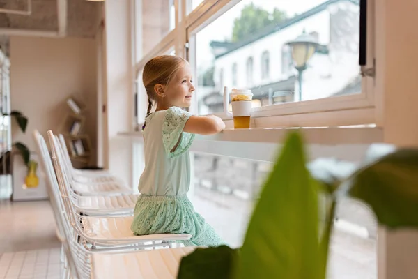 Carino piccola ragazza caucasica di sette anni indossa abbigliamento casual e bere tè alla frutta al caffè — Foto Stock