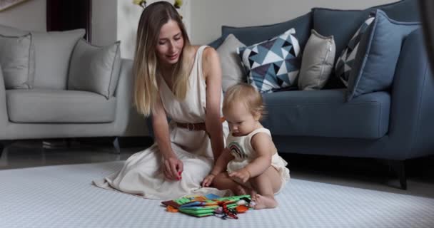 Giovane madre caucasica o tata che gioca con il bambino carino di un anno, imparando a leggere il libro di sviluppo per bambini piccoli. Due generazioni di famiglia che trascorrono del tempo insieme e si godono la vita sul pavimento di casa — Video Stock