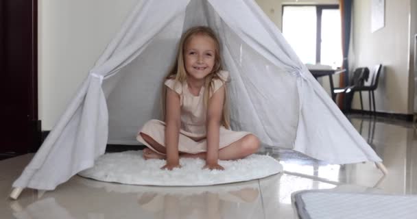 Upřímný portrét docela roztomilé malé kavkazské dívky s blond vlasy sedí doma ve vigvamu. Krásné dítě sedm let s usměvavou tváří při pohledu na kameru v pomalém pohybu. — Stock video