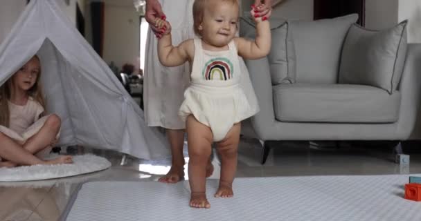 Mignon caucasien Bébé fille aux cheveux blonds et aux yeux bleus d'un an apprenant à marcher. Infantile faisant les premiers pas à la maison. Mère aidant l'enfant à apprendre à partir. Joyeux nouveau concept de vie. Petits pieds — Video