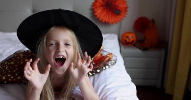 Cadı elbisesi giymiş, şapkalı, sarı saçlı sevimli beyaz kız Coronavirus covid-19 salgını ve karantinası sırasında evde eğleniyor ve Cadılar Bayramı 'nı kutluyor. Yavaş çekim