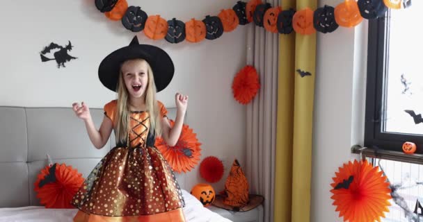 Petite fille blanche mignonne aux cheveux blonds de sept ans en robe de sorcière et chapeau s'amuser et célébrer Halloween à la maison pendant la pandémie et la quarantaine de Coronavirus covid-19. Mouvement lent — Video