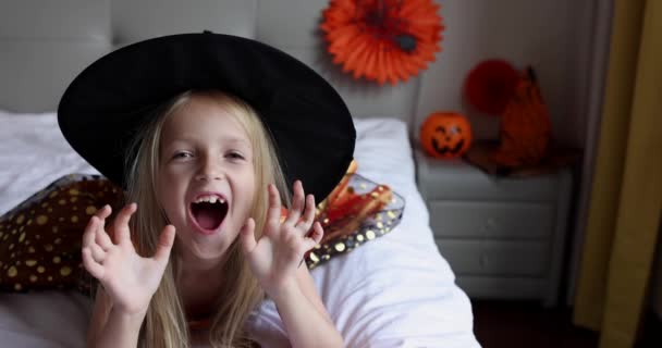 Petite fille blanche mignonne aux cheveux blonds de sept ans en robe de sorcière et chapeau s'amuser et célébrer Halloween à la maison pendant la pandémie et la quarantaine de Coronavirus covid-19. Mouvement lent — Video