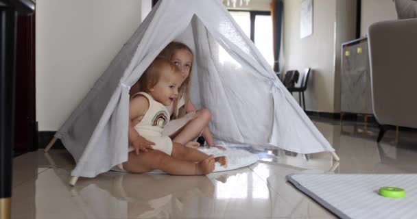 Två söta små kaukasiska barn syskon leker tillsammans i wigwam på golvet hemma. Koncept för barns utveckling. Spädbarn flicka med blont hår leker med äldre syster. Långsamma rörelser 4k — Stockvideo