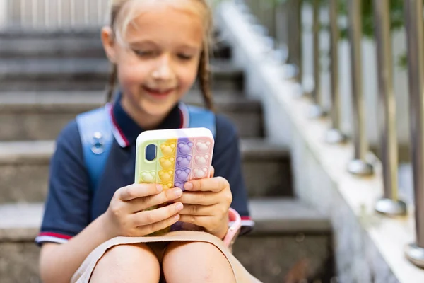 Lilla kaukasiska flicka leker med mobiltelefon i ny pop det leksak fall. Utveckling av finmotorik, Push bubbla fidget sensorisk leksak - tvättbar och återanvändbar kisel stress lättnad leksak. Anti — Stockfoto