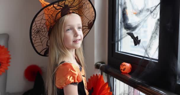 Linda niña caucásica con el pelo rubio de siete años de edad en vestido de bruja y sombrero divertirse y celebrar Halloween en casa durante Coronavirus covid-19 pandemia y cuarentena. Movimiento lento — Vídeo de stock