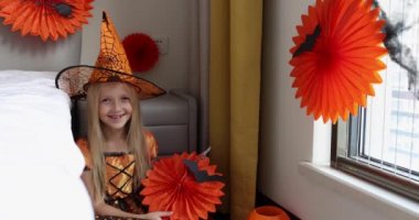 Cadı elbisesi giymiş, şapkalı, sarı saçlı sevimli beyaz kız Coronavirus covid-19 salgını ve karantinası sırasında evde eğleniyor ve Cadılar Bayramı 'nı kutluyor. Yavaş çekim