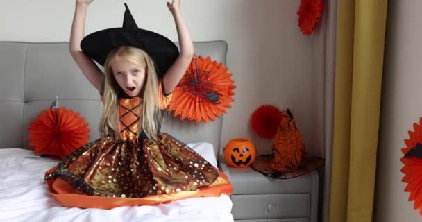 Linda niña caucásica con el pelo rubio de siete años de edad en vestido de bruja y sombrero divertirse y celebrar Halloween en casa durante Coronavirus covid-19 pandemia y cuarentena. Movimiento lento — Vídeo de stock