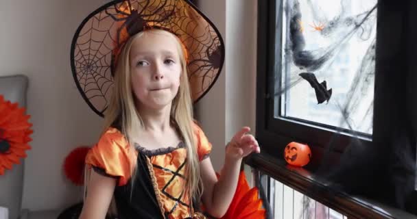 Schattig blank klein meisje met blond haar zeven jaar oud in heksenjurk en hoed plezier hebben en Halloween vieren thuis tijdens Coronavirus covid-19 pandemie en quarantaine. Langzame beweging — Stockvideo