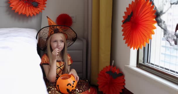 Мила кавказька дівчинка з білявим волоссям у відьминому вбранні і капелюсі, що веселиться і святкує Хелловін вдома під час коронавірусної конід-19 пандемії і карантину. Повільний рух — стокове відео