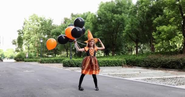 Linda niña caucásica con el pelo rubio de siete años de edad en traje de bruja con sombrero y vestido naranja negro posando con un montón de globos de Halloween en la calle. Concepto de vacaciones. Movimiento lento. — Vídeos de Stock