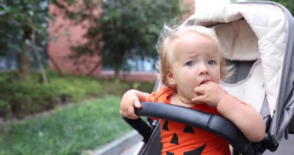 Linda niña caucásica con cabello rubio de un año de edad en traje naranja con cara de calabaza para Halloween sentado en el carro al aire libre. Concepto de vacaciones. Movimiento lento. — Vídeos de Stock