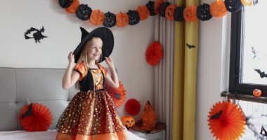 Cadı elbisesi giymiş, şapkalı, sarı saçlı sevimli beyaz kız Coronavirus covid-19 salgını ve karantinası sırasında evde eğleniyor ve Cadılar Bayramı 'nı kutluyor. Yavaş çekim