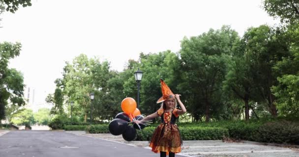 Söt liten kaukasiska flicka med blont hår sju år gammal i dräkt av häxa med hatt och svart orange klänning poserar med gäng halloween ballonger på gatan. Semesterkoncept. Långsamma rörelser. — Stockvideo