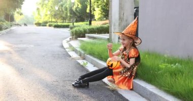 Yedi yaşında, sarı saçlı, şapkalı, siyah turuncu elbiseli, caddede cadılar bayramını kutlayan bir cadı kostümü giyen sevimli, beyaz bir kız. Tatil konsepti. Yavaş çekim.