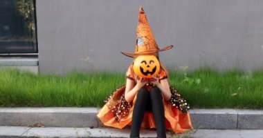 Yedi yaşında, sarı saçlı, şapkalı, siyah turuncu elbiseli, caddede cadılar bayramını kutlayan bir cadı kostümü giyen sevimli, beyaz bir kız. Tatil konsepti. Yavaş çekim.