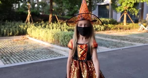 Söt liten kaukasiska flicka med blont hår sju år gammal i dräkt av häxa med hatt och svart orange klänning firar Halloween utomhus på gatan. Semesterkoncept. Långsamma rörelser. — Stockvideo