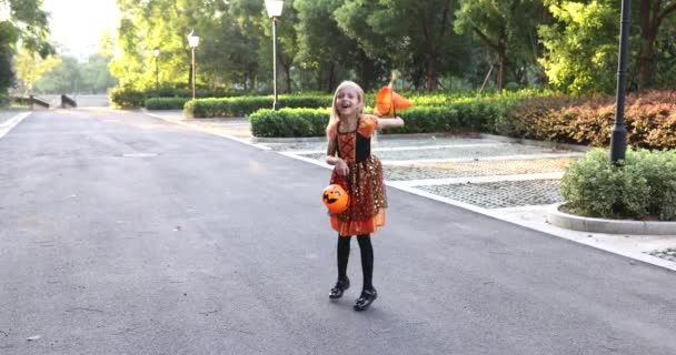 Schattig klein blank meisje met blond haar zeven jaar oud in kostuum van heks met hoed en zwarte oranje jurk vieren Halloween buiten op straat. Vakantie concept. Langzame beweging. — Stockvideo