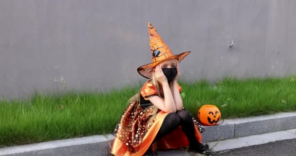 Linda niña caucásica con el pelo rubio de siete años de edad en traje de bruja con sombrero y vestido naranja negro celebrando Halloween al aire libre en la calle. Concepto de vacaciones. Movimiento lento. — Vídeo de stock