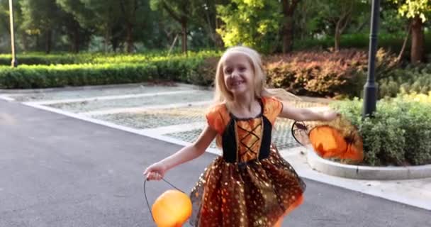 Linda niña caucásica con el pelo rubio de siete años de edad en traje de bruja con sombrero y vestido naranja negro celebrando Halloween al aire libre en la calle. Concepto de vacaciones. Movimiento lento. — Vídeos de Stock