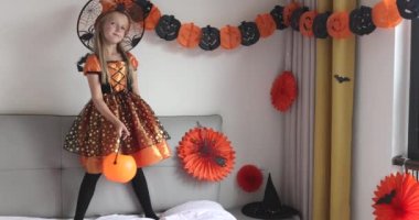 Cadı elbisesi giymiş, şapkalı, sarı saçlı sevimli beyaz kız Coronavirus covid-19 salgını ve karantinası sırasında evde eğleniyor ve Cadılar Bayramı 'nı kutluyor. Yavaş çekim