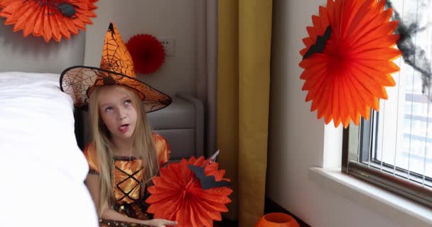 Linda niña caucásica con el pelo rubio de siete años de edad en vestido de bruja y sombrero divertirse y celebrar Halloween en casa durante Coronavirus covid-19 pandemia y cuarentena. Movimiento lento — Vídeo de stock