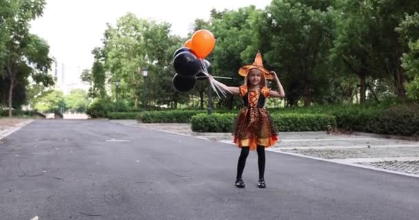 Roztomilé malé běloška dívka s blond vlasy sedm let v kostýmu čarodějnice s kloboukem a černě oranžové šaty pózovat s partou halloween balónky na ulici. Koncept dovolené. Zpomalený pohyb. — Stock video