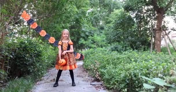 Linda niña caucásica con el pelo rubio de siete años de edad en traje de bruja con vestido naranja negro celebrando Halloween al aire libre en la calle. Concepto de vacaciones. Movimiento lento. — Vídeos de Stock