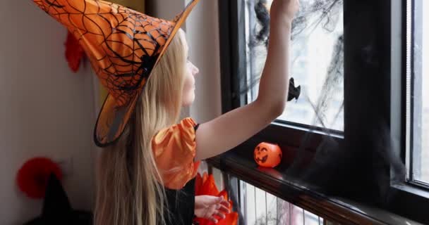 Linda niña caucásica con el pelo rubio de siete años de edad en vestido de bruja y sombrero divertirse y celebrar Halloween en casa durante Coronavirus covid-19 pandemia y cuarentena. Movimiento lento — Vídeo de stock