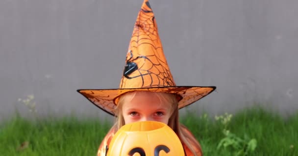 Linda niña caucásica con el pelo rubio de siete años de edad en traje de bruja con sombrero y vestido naranja negro celebrando Halloween al aire libre en la calle. Concepto de vacaciones. Movimiento lento. — Vídeos de Stock